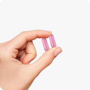 For Conceive Plus Women's Fertility Support and Ovulation Support Bundle, a close-up of two pink fertility capsules of Women's Fertility Support held delicately between thumb and forefinger, highlighting capsule size and color on a clean, white background.